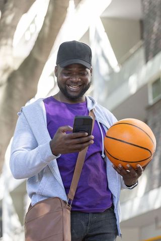 African american man holding basketball and checking smartphone while smiling outdoors