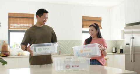 Couple sorting recycling in modern kitchen holding labeled clear bins for paper plastic glass