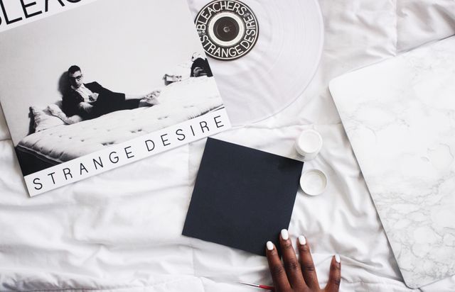 Relaxing scene illustrating modern minimalist home lifestyle. A person holding a blank black card is beside a vinyl record on white bedsheets, along with marble-patterned laptop. Perfect for blog posts or articles on music, minimalistic decoration, modern lifestyle, or cozy home settings.