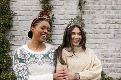 Two friends standing and smiling outdoors wearing festive tiaras and cozy sweaters