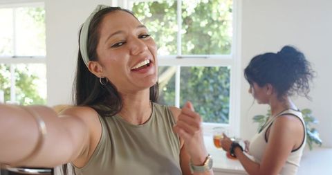Smiling woman taking selfie in sunlit kitchen while friend pours tea, morning routine