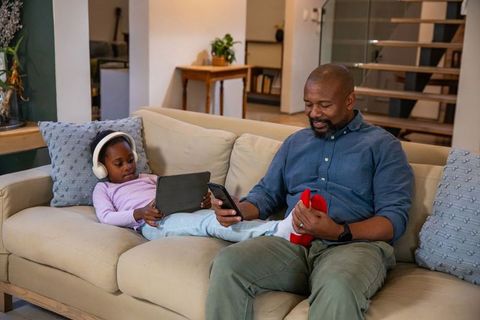 African american father helping daughter while using phone and tablet on cozy home sofa