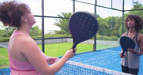 Women playing padel on blue court holding black rackets at net for outdoor training