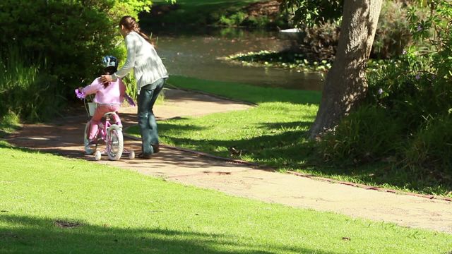 This stock photo captures a mother guiding her daughter while she rides a bike in a lovely park. Set on a sunny day, the scene highlights family bonding, childhood learning experiences, and outdoor activities. Ideal for use in topics related to parenting, family fun, healthy lifestyles, and outdoor recreation.