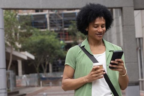 African american commuter standing with takeaway coffee checking smartphone outdoors