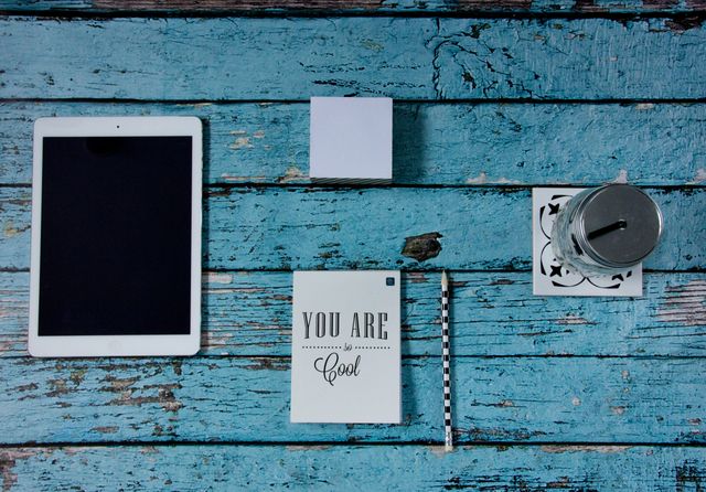 Creative workspace with electronic tablet, note card reading 'You Are Cool', and striped pencil on rustic blue wooden background. Ideal for use in design blogs, productivity articles, or inspirational social media posts highlighting neat and organized workspaces.