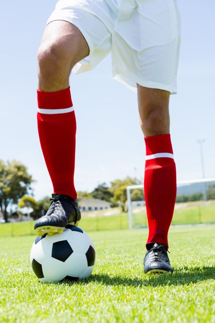 Football player standing with one foot on a soccer ball in a stadium. The player is wearing red socks and white shorts, with a clear blue sky in the background. Ideal for use in sports advertisements, athletic promotions, soccer training materials, and recreational activity brochures.