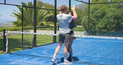 High-fiving couple playing padel on vibrant blue court outdoors, sunlit lifestyle sport