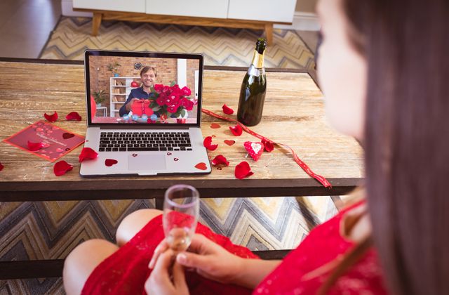 Caucasian mid adult man and biracial girlfriend celebrate Valentine's Day over an online video call. Background features romantic decorations, gifts, and champagne. Suitable for themes of virtual relationships, love during the pandemic, digital celebrations, and long-distance dating.