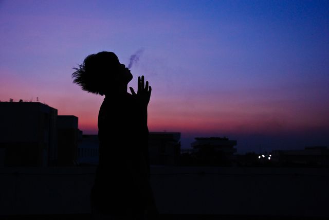 Silhouette of person releasing smoke into the vibrant sunset sky on a rooftop in the city. The article showcases relaxation and contemplation, making it suitable for topics related to urban lifestyle, evening relaxation, meditative practices, and mental health themes. Ideal for blogs, social media posts, urban studies, and lifestyle magazines.
