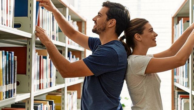 Young couple browsing books on library shelves, collaborating, smiling in natural daylight