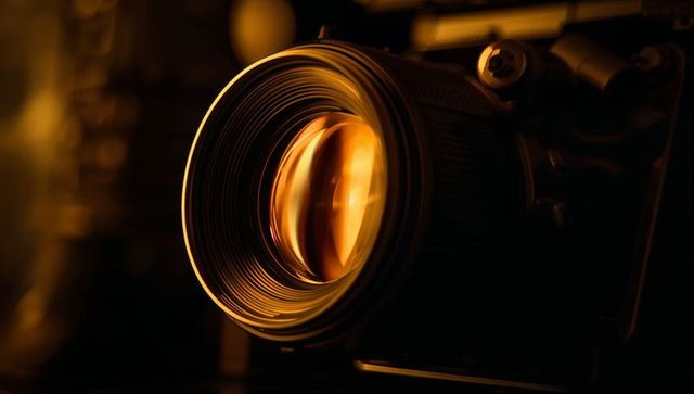 Glowing amber camera lens close-up featuring focus ring, metal details and studio bokeh