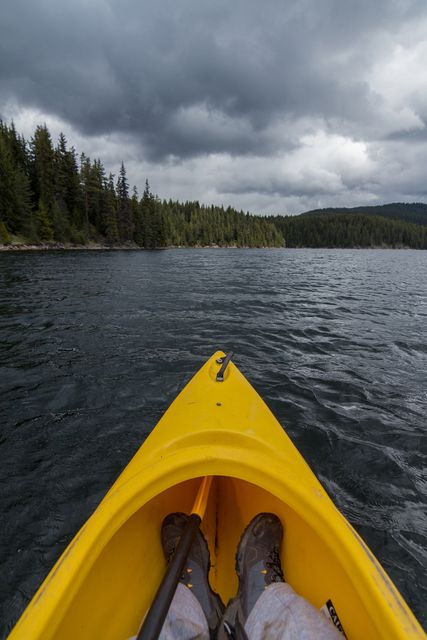 Person kayaking in yellow kayak on a lake surrounded by forest under a cloudy sky. Suitable for articles or advertisements about outdoor activities, leisure, travel destinations, or kayaking experiences.