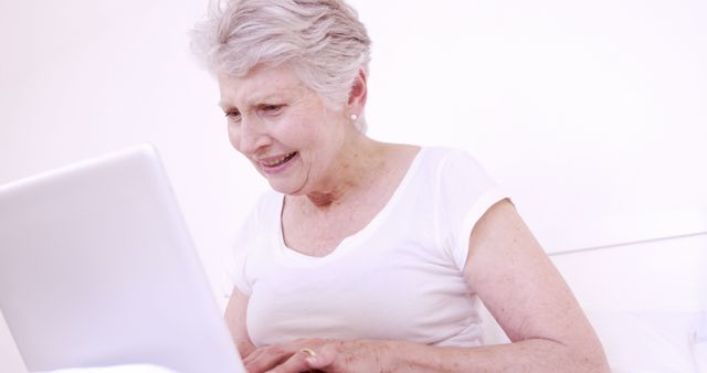 An elderly woman with grey hair and casual clothing is sitting in a white room and actively using a laptop. She is smiling and appears to be engaged with the content on the screen. Ideal for topics on seniors using technology, online learning for older adults, health and wellness in aging, and promoting tech-savviness among the elderly.