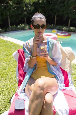 Mid adult woman sipping lemon water in yellow swimsuit by backyard pool lounger
