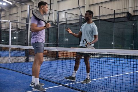 Two men chatting across net on indoor padel court holding paddles wearing athletic gear
