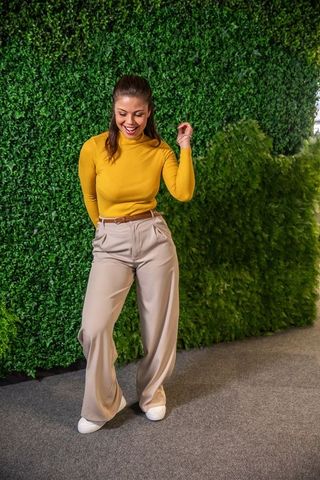 Smiling woman standing by green plant wall wearing mustard turtleneck and wide leg trousers