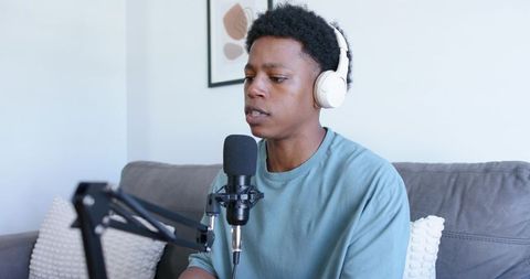 Young podcaster recording at home with white headphones and microphone on grey sofa