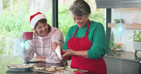 Two women decorating christmas cookies and laughing in sunlit kitchen, festive baking