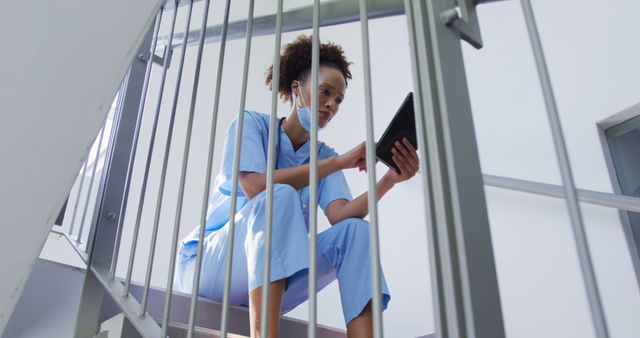 Biracial female doctor wearing face mask sitting on stairs in hospital using tablet. medicine, health and healthcare services during coronavirus covid 19 pandemic.