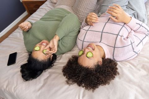 Asian and african american friends lying on bed with cucumber eye masks relaxing