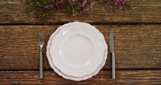 An empty white plate is flanked by a fork on the left and a knife on the right, set on a rustic wooden table adorned with pink flowers, with copy space. The arrangement suggests a natural and inviting setting for a meal, emphasizing simplicity and elegance.