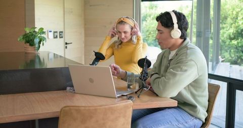 Podcast duo recording at kitchen table with laptop and microphones in modern home studio