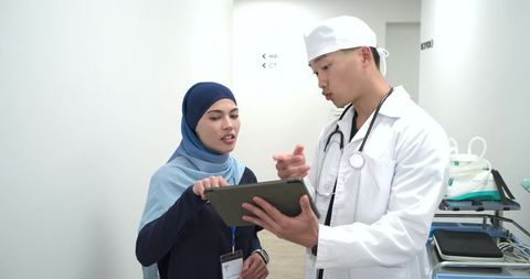 Doctor consulting hijab-wearing colleague reviewing patient data on tablet in clinic corridor