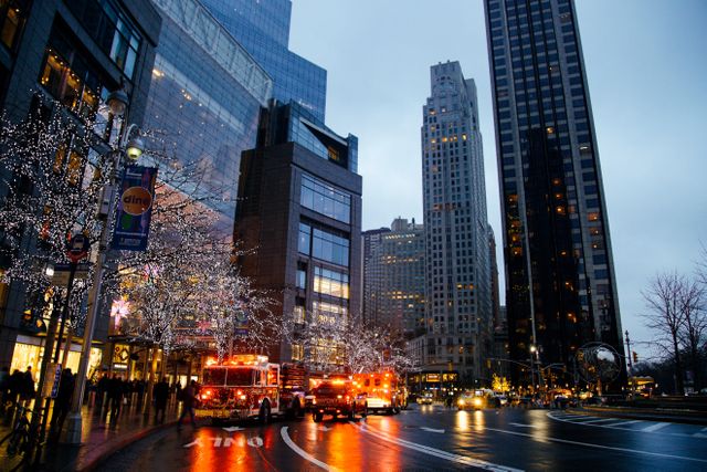 Street scene in a city during dusk with fire trucks and illuminated trees. Skyscrapers and buildings create an urban setting, with emergency response vehicles providing an element of urgency. Useful for themes involving city life, emergency services, urban areas, night scenes, and evening cityscapes.