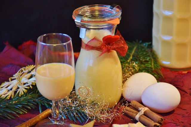Christmas eggnog displayed in a cork-sealed jar adorned with a ribbon alongside a filled glass. Surrounding elements include eggs, cinnamon sticks, Christmas tree branches, and twine, creating a festive holiday atmosphere. Ideal for use in holiday promotions, celebration content, and festive recipes.