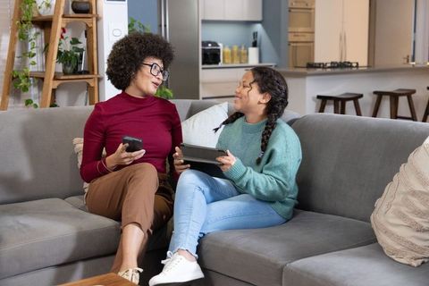 African-american mother and daughter bonding on couch with tablet and phone in modern home