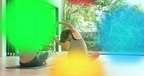 Two women practicing seated side-bend yoga at home with sunlit garden and wood floor