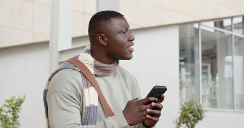 African american man using smartphone standing beside glass facade wearing knit scarf