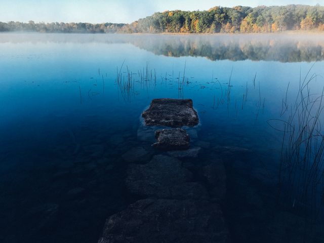 This image captures a tranquil and serene morning at a misty lake with a stone path leading to the foggy horizon. Surrounding autumn trees and peaceful blue water add to the stillness. Ideal for use in nature-themed blogs, meditation and relaxation content, inspirational posters, or as a backdrop for calming music playlists.