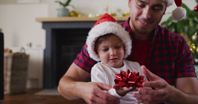 Father and young son wearing Santa hats unwrapping a gift at home. The father is assisting the child with the present, creating a heartwarming family moment. Perfect for use in holiday greetings, family bonding promotions, Christmas advertisements, and festive season campaigns emphasizing family joy and togetherness.