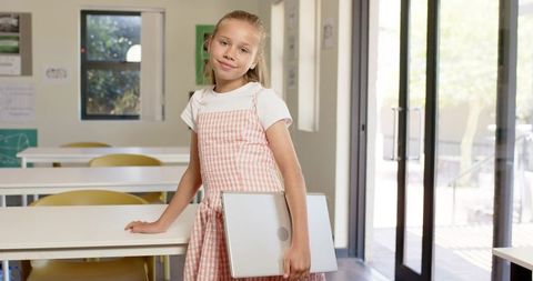 Preteen student holding laptop in bright classroom wearing pink pinafore and white tee