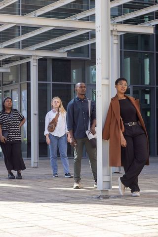 Diverse professionals walking through urban plaza with woman leaning on column wearing brown coat, g