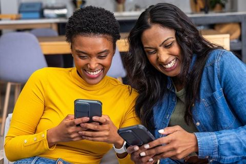 Smiling diverse women checking phones on sofa, casual coworking and friendship moment