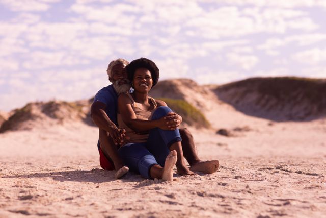 African american couple looking away while sitting on sandy beach against cloudy sky in summer. afro hair, beard, unaltered, love, togetherness, retirement, enjoyment and holiday concept.