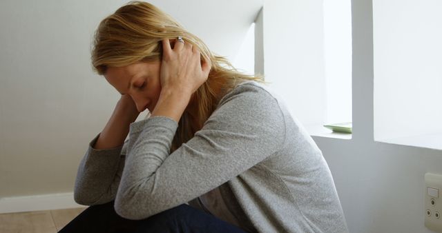 This stock photo depicts a woman in a gray sweater sitting on the floor, holding her head in her hands, visibly stressed and worried. This image is ideal for articles or advertisements related to mental health, emotional well-being, anxiety, depression, and personal crises. It can be used by blogs, therapy websites, health magazines, and other platforms discussing psychological topics.