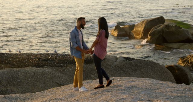 Diverse couple holding hands on rocky beach at sunset. Both have dark hair, woman wearing a pink top and jeans, the man in a blue shirt and yellow pants