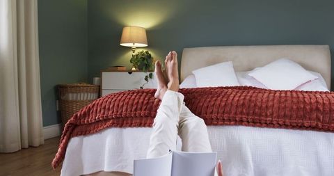 Relaxed Woman Reading Book in Cozy Bedroom with Rust Red Bedding and Ambient Lighting