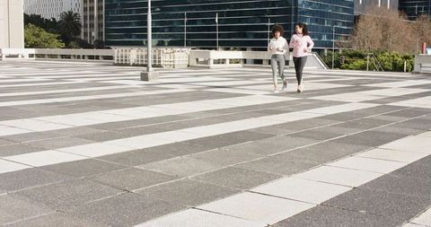 Two female friends jogging on striped urban plaza with glass office tower backdrop