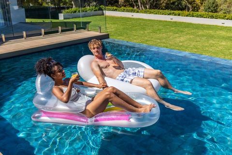 Relaxing interracial couple lounging on inflatable pool floats sipping drinks in backyard