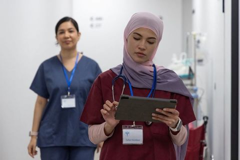 Muslim nurse reviewing tablet while colleague walking hospital corridor, clinical teamwork