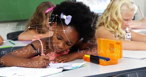 Young girl writing in workbook while focusing on math with classroom stationery and classmates