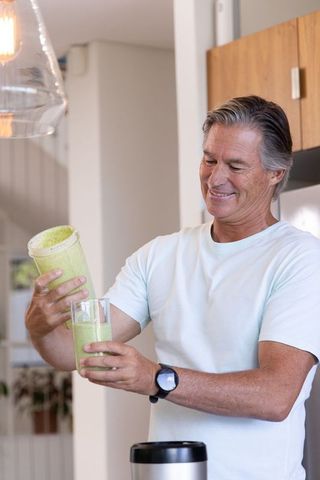 Mature man pouring green smoothie into glass in bright minimalist home kitchen