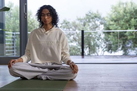 Non-binary adult meditating on green mat by glass doors wearing cream hoodie and gray sweatpants
