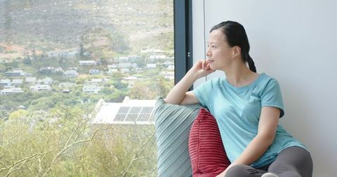 Asian woman sitting and gazing out window from cozy couch with hillside view and solar panels
