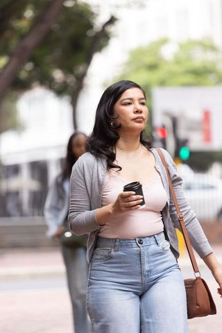 Urban commuter walking with takeaway coffee and leather shoulder bag in casual outfit